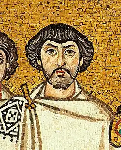 A close up photograph of a mosaic of a person painted on a golden coloured background. The person has a white cloth covering up his upper body till the neck, an embroidered pattern on his right shoulder, wavy hair, a stubble beard and a moustache
